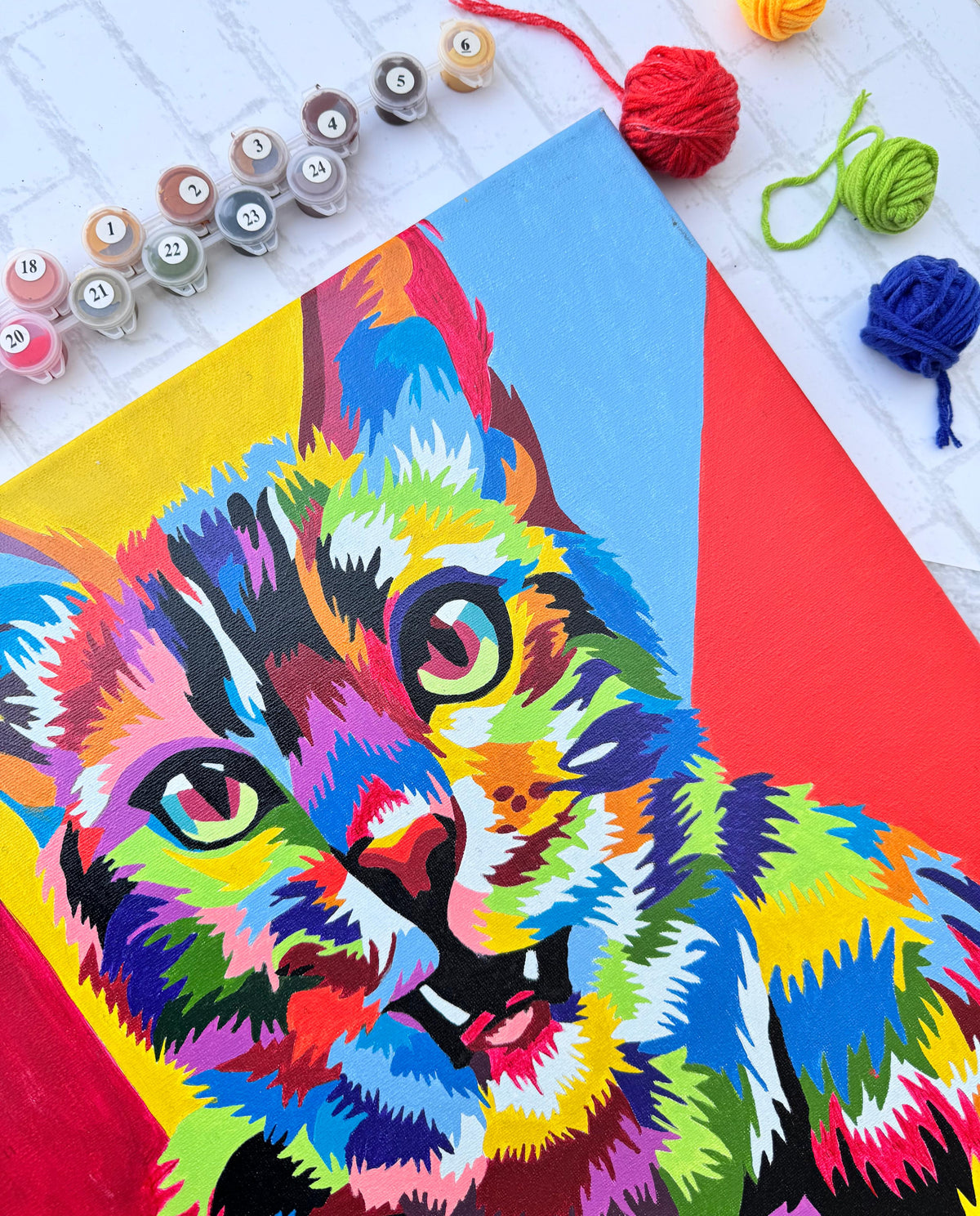 Lienzo de gato multicolor para pintar por números con pinceles y botes de pintura junto a ovillos de lana roja, verde y azul.