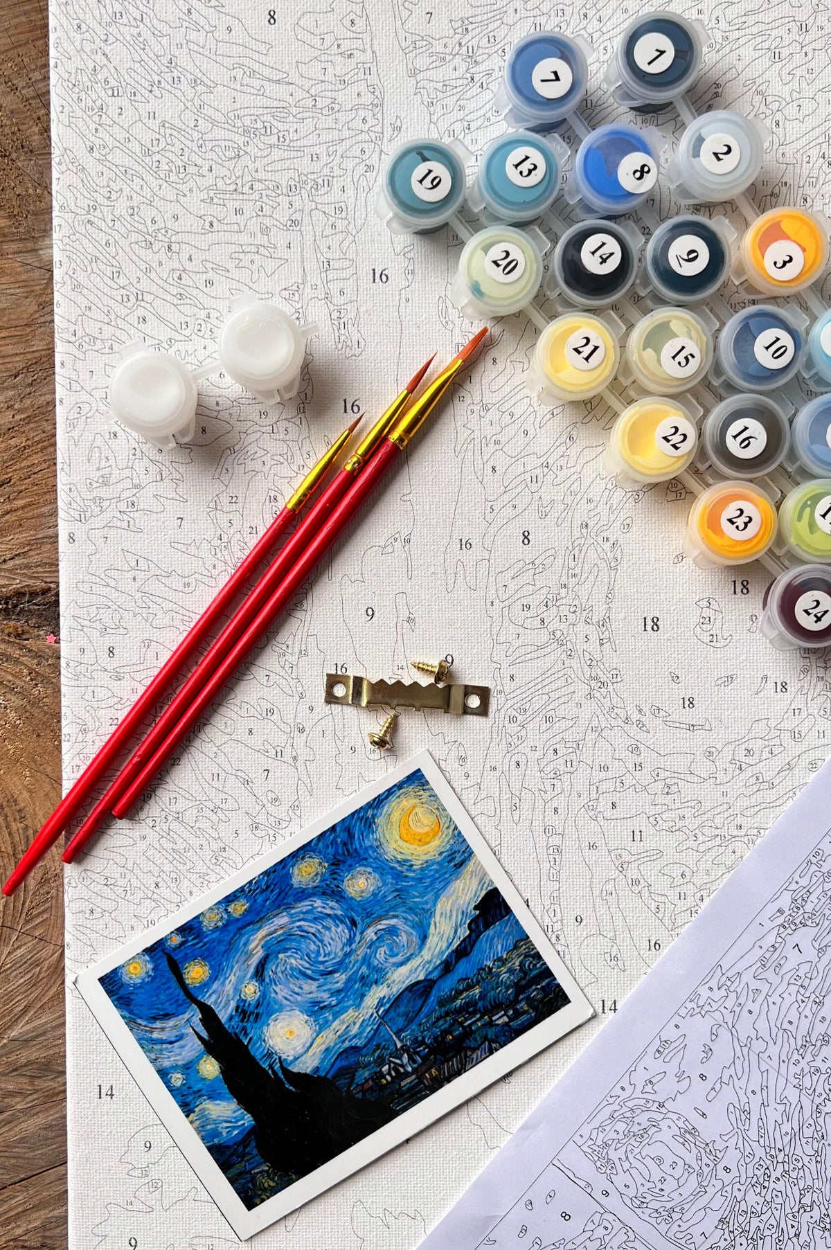 Kit de pinturas por números Noche Estrellada con pinceles rojos y pequeños botes de pintura numerados y plantilla en fondo.