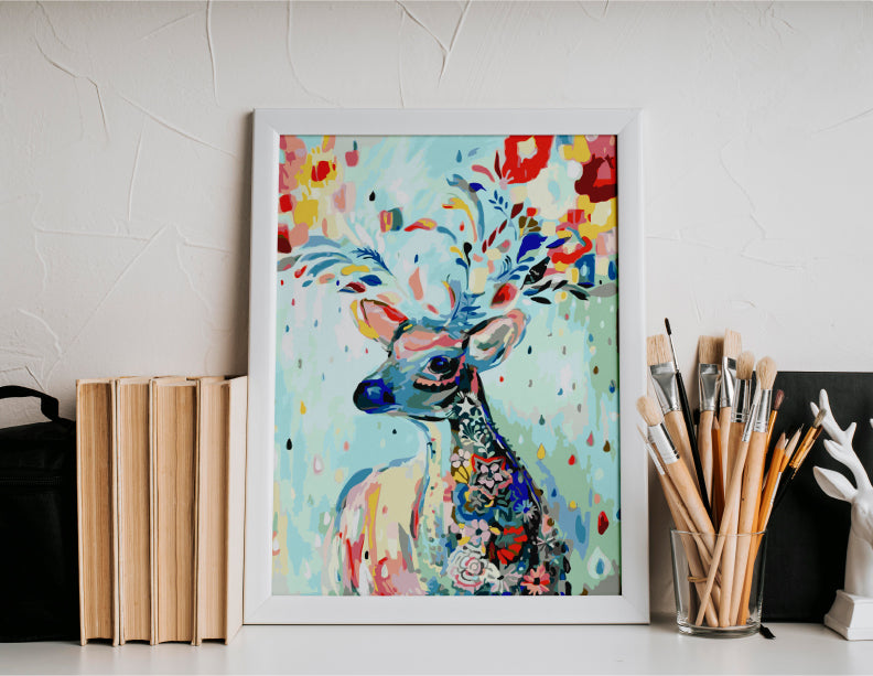 Cuadro de reno colorido pintado con estilo artístico abstracto en marco blanco sobre mesa junto a pinceles y libros.