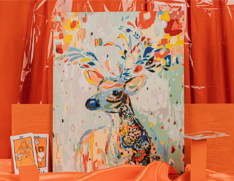 Lienzo de reno colorido con pintura por números, acompañado de pinceles y paleta naranja.